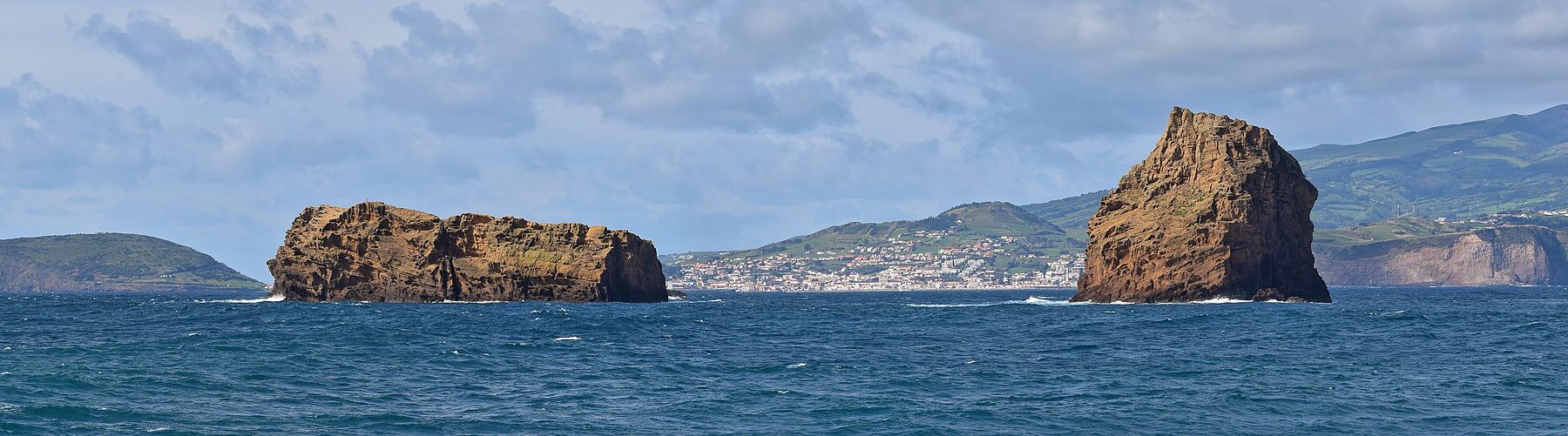 Panorama des deux îlots de tuf ocre de Madalena sur une mer bleu sombre, l'Ilhéu Deitado long et plat à gauche et l'Ilhéu em Pé dressé à droite, avec la ville blanche de Horta et les collines vertes de Faial au fond