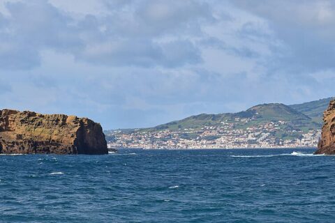 Panorama des deux îlots de tuf ocre de Madalena sur une mer bleu sombre, l'Ilhéu Deitado long et plat à gauche et l'Ilhéu em Pé dressé à droite, avec la ville blanche de Horta et les collines vertes de Faial au fond