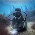 Un plongeur en combinaison noire, allongé sur un fond de sable entre deux rochers couverts d'algues brunes, écrit sur une ardoise blanche, des silhouettes de laminaires se découpant dans le bleu derrière lui.