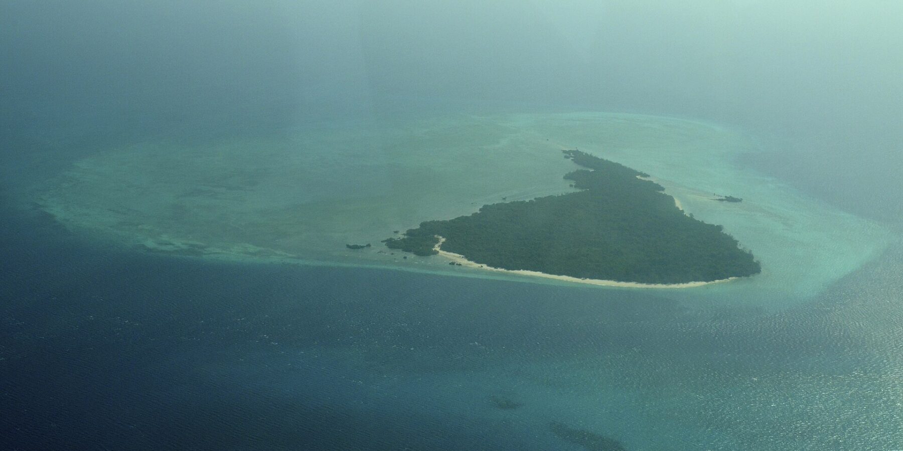 L'île de Misali vue d'avion, triangle de forêt bordé d'une plage blanche, posé sur un platier turquoise au milieu d'une mer bleu sombre, dans la brume