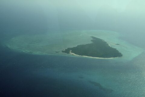 L'île de Misali vue d'avion, triangle de forêt bordé d'une plage blanche, posé sur un platier turquoise au milieu d'une mer bleu sombre, dans la brume