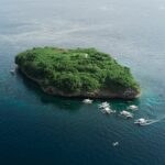 Vue aérienne de l'île de Pescador, un rocher couvert de végétation verte avec son phare blanc, entouré d'une dizaine de bancas de plongée sur une mer bleu foncé