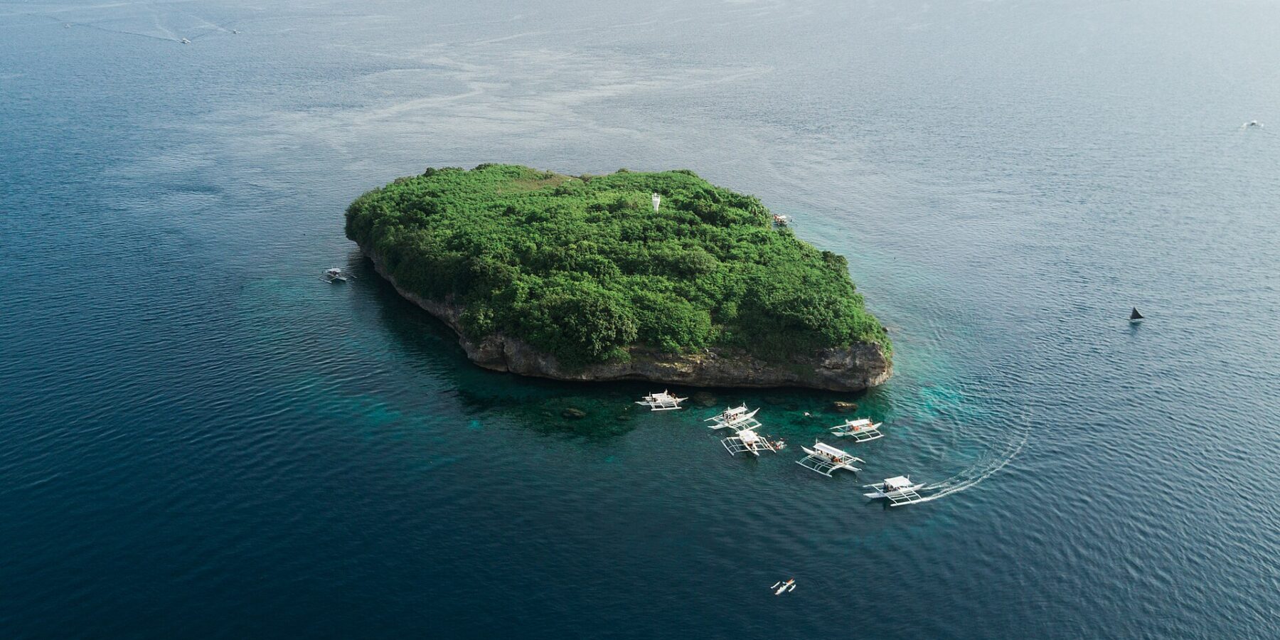 Vue aérienne de l'île de Pescador, un rocher couvert de végétation verte avec son phare blanc, entouré d'une dizaine de bancas de plongée sur une mer bleu foncé