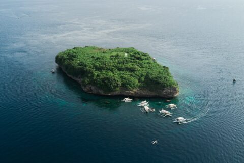 Vue aérienne de l'île de Pescador, un rocher couvert de végétation verte avec son phare blanc, entouré d'une dizaine de bancas de plongée sur une mer bleu foncé