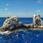 Roca Partida vue de drone : un rocher blanchi par les oiseaux, fendu en deux pics, battu par une mer bleu foncé sous un ciel bleu parsemé de nuages