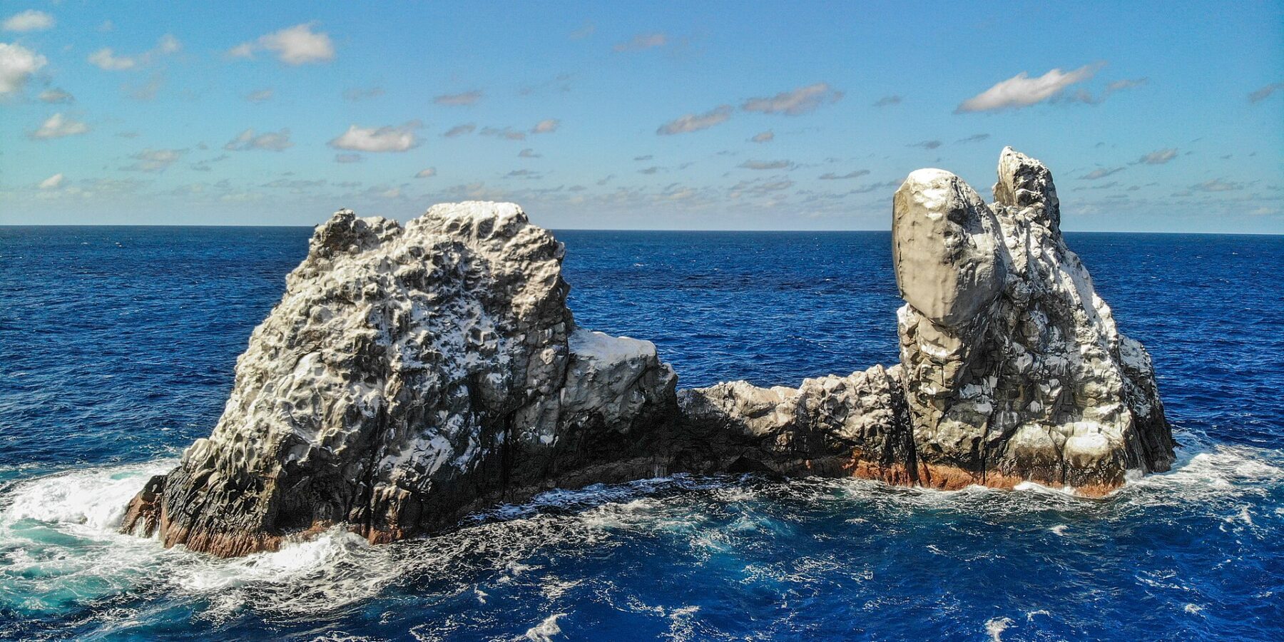 Roca Partida vue de drone : un rocher blanchi par les oiseaux, fendu en deux pics, battu par une mer bleu foncé sous un ciel bleu parsemé de nuages