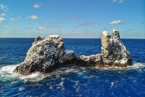 Roca Partida vue de drone : un rocher blanchi par les oiseaux, fendu en deux pics, battu par une mer bleu foncé sous un ciel bleu parsemé de nuages