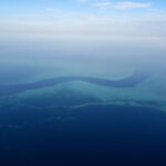 Vue d'avion d'un chenal bleu sombre en forme de S qui serpente entre les platiers turquoise de la barrière de corail, quelques petits bateaux blancs dans la passe