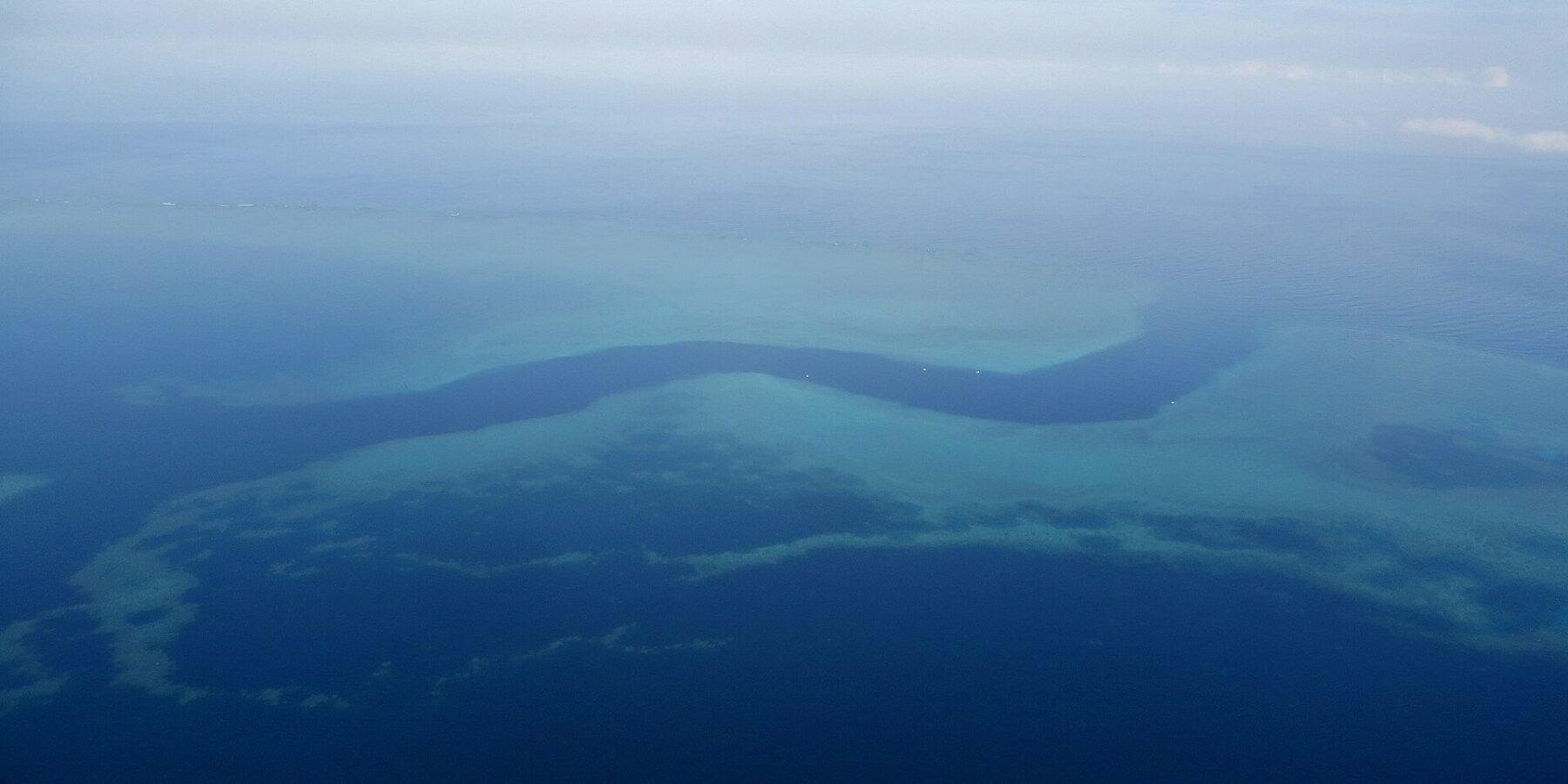 Vue d'avion d'un chenal bleu sombre en forme de S qui serpente entre les platiers turquoise de la barrière de corail, quelques petits bateaux blancs dans la passe