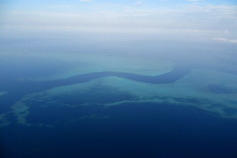 Vue d'avion d'un chenal bleu sombre en forme de S qui serpente entre les platiers turquoise de la barrière de corail, quelques petits bateaux blancs dans la passe