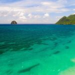 Vue aérienne de la longue plage du Diamant bordée d'eau turquoise, le Rocher du Diamant posé au large et le morne Larcher, une colline verte, à droite
