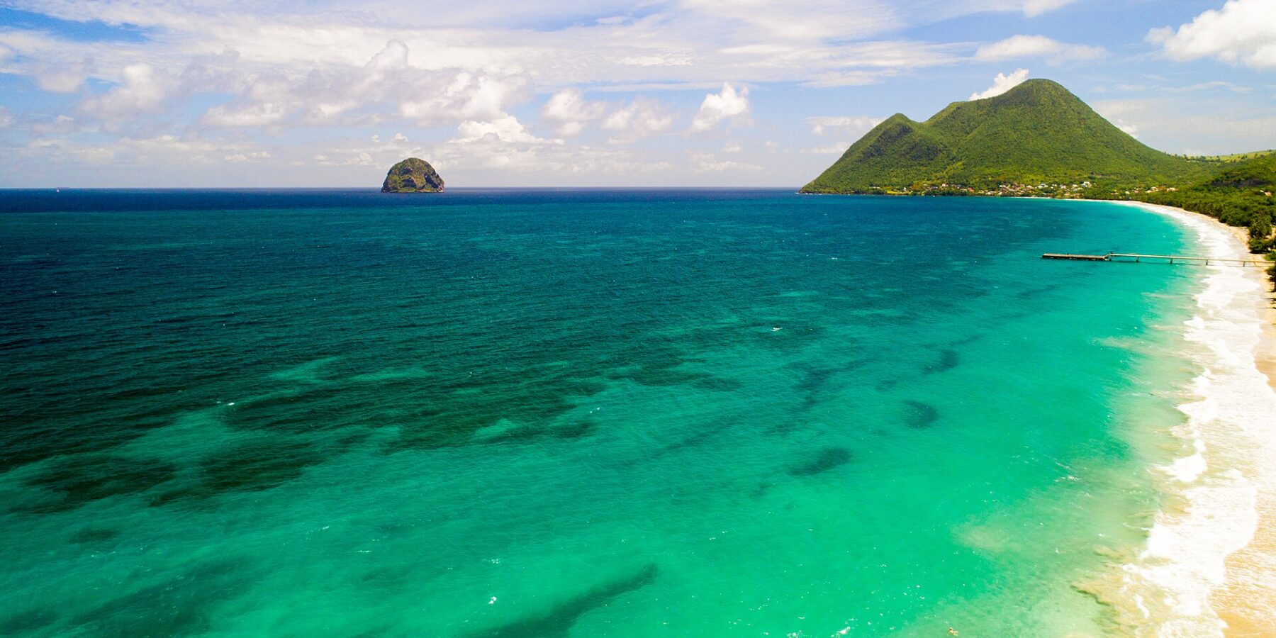 Vue aérienne de la longue plage du Diamant bordée d'eau turquoise, le Rocher du Diamant posé au large et le morne Larcher, une colline verte, à droite