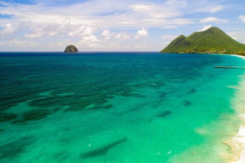 Vue aérienne de la longue plage du Diamant bordée d'eau turquoise, le Rocher du Diamant posé au large et le morne Larcher, une colline verte, à droite