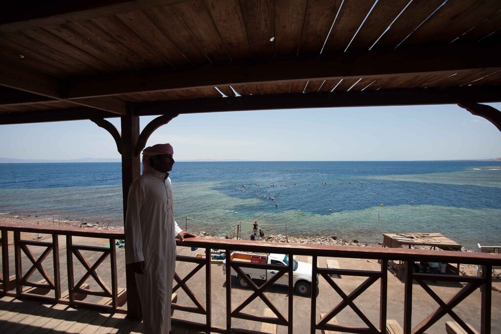 Depuis une terrasse en bois, un homme en galabeya regarde la vasque du Blue Hole, bordée de récif clair, où nagent des baigneurs