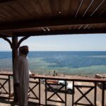Depuis une terrasse en bois, un homme en galabeya regarde la vasque du Blue Hole, bordée de récif clair, où nagent des baigneurs