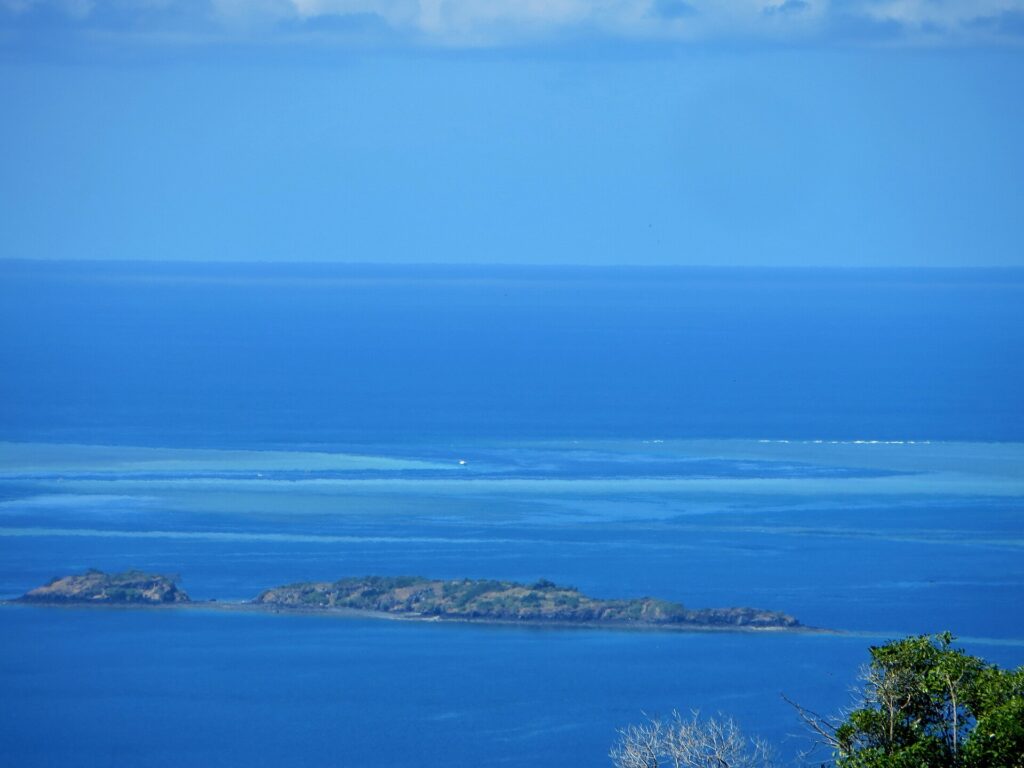 Vue plongeante sur le lagon bleu, deux îlots rocheux au premier plan et, au loin, les platiers clairs de la barrière de corail où un bateau blanc stationne dans la passe