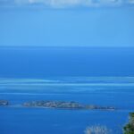Vue plongeante sur le lagon bleu, deux îlots rocheux au premier plan et, au loin, les platiers clairs de la barrière de corail où un bateau blanc stationne dans la passe